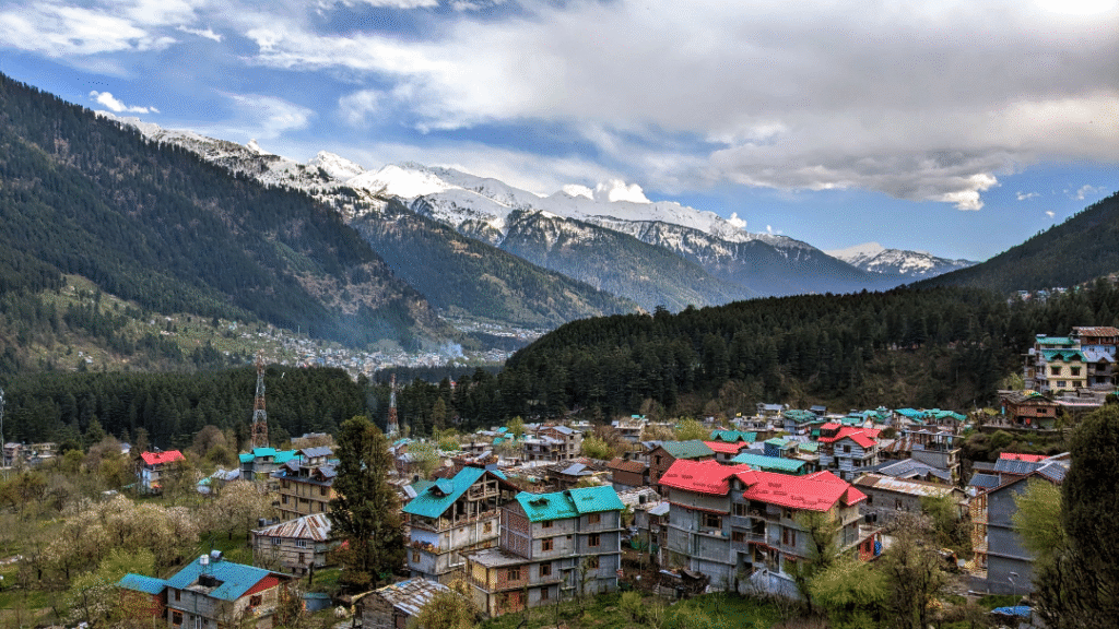A panoramic view of Manali, Himachal Pradesh, featuring snow-capped peaks, dense pine forests, and a peaceful valley below under a bright blue sky.
