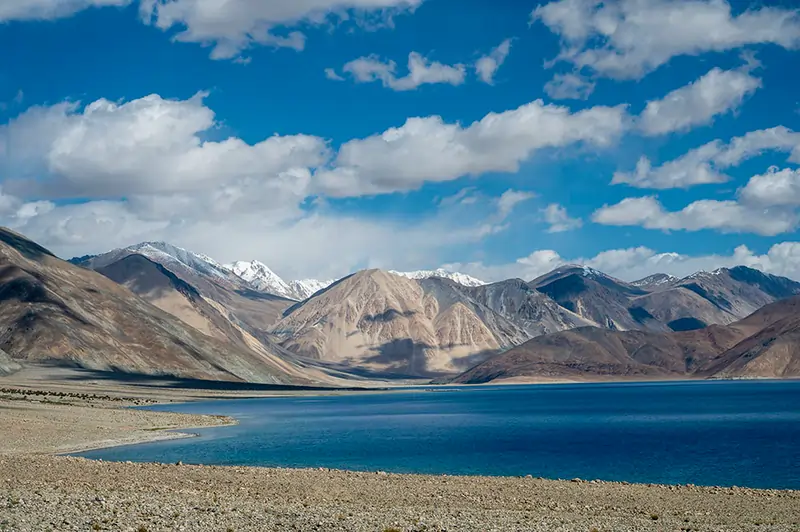  A dramatic landscape of Leh-Ladakh featuring barren mountains, a winding road, and a clear blue sky with scattered clouds, highlighting the region’s rugged and serene beauty.