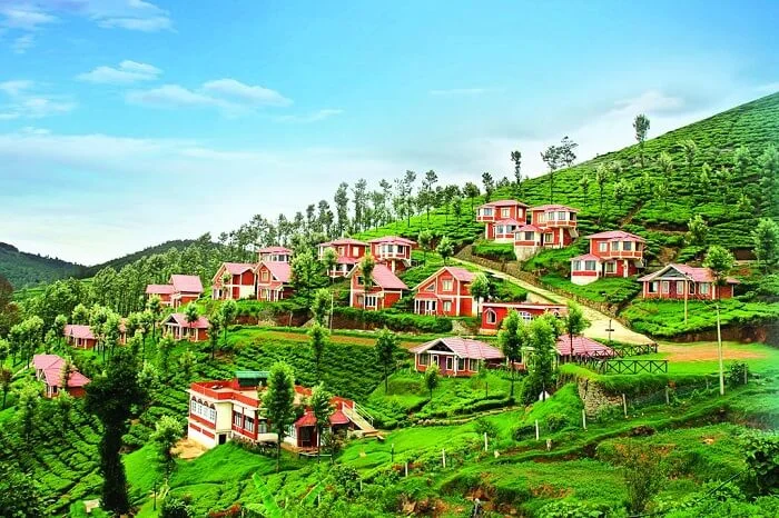  A scenic view of Ooty, Tamil Nadu, featuring rolling hills covered in lush greenery, colorful houses scattered across the slopes, and the iconic Nilgiri Mountain Railway train curving through the landscape.