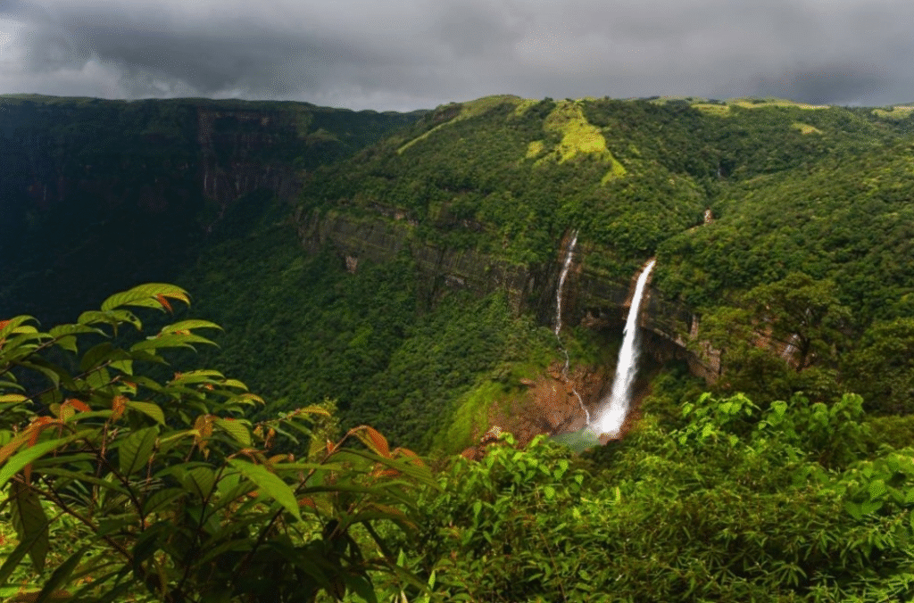 A misty landscape of Shillong, Meghalaya, with rolling green hills, cascading waterfalls, and scattered traditional houses nestled among pine trees under a cloudy sky