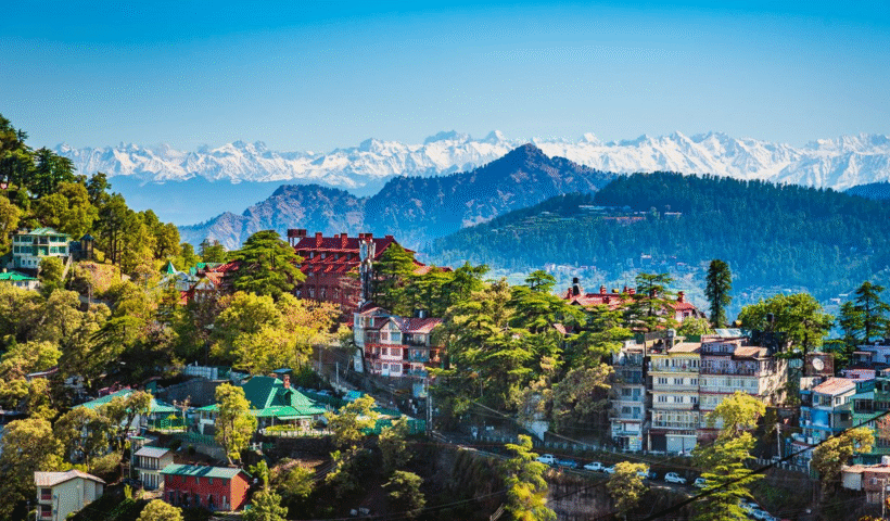 A scenic view of Shimla, Himachal Pradesh, showing colonial-style buildings nestled among lush green hills and mist-covered pine forests under a soft, cloudy sky.
