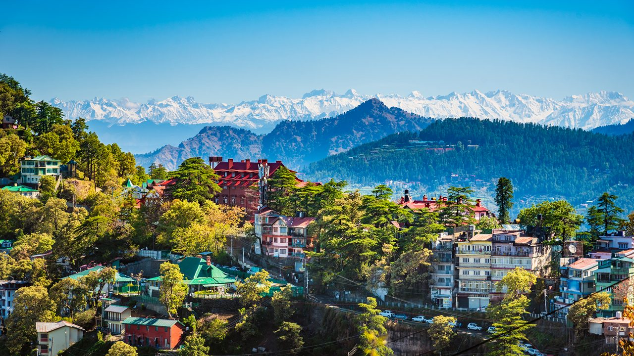 A scenic view of Shimla, Himachal Pradesh, showing colonial-style buildings nestled among lush green hills and mist-covered pine forests under a soft, cloudy sky.